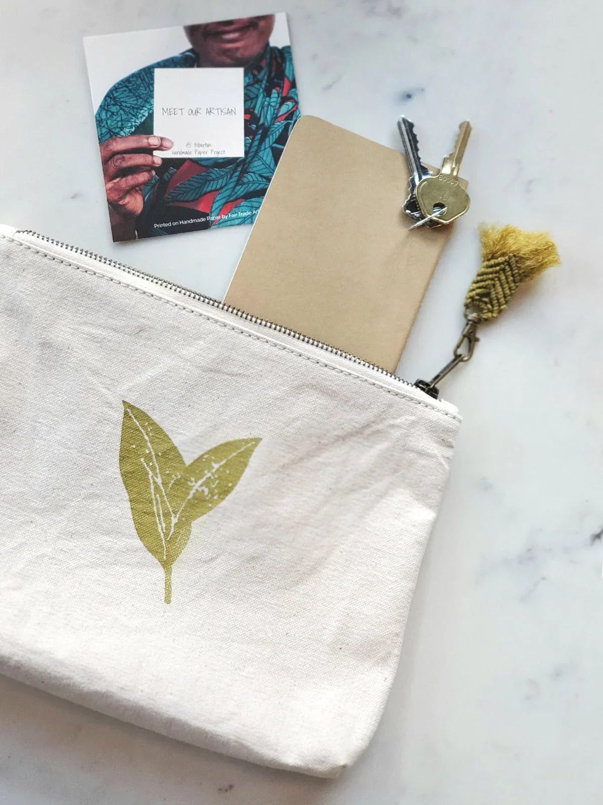 Eco-friendly canvas pouch with green leaf print, keys, notebook, and artisan card on marble
