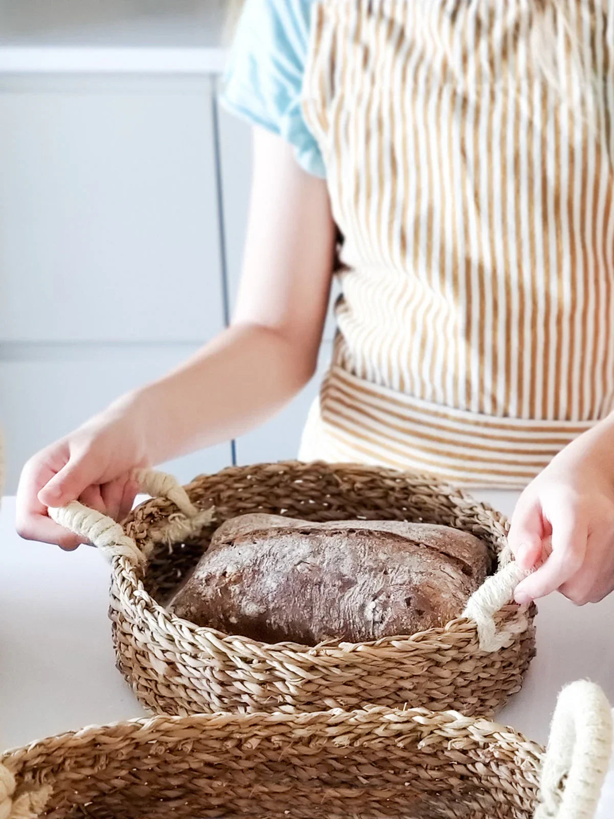Person holding round woven savar bread basket with loaf of rustic bread on white table