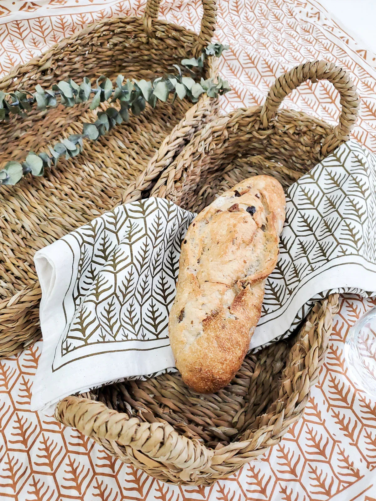 Set of two natural woven bread baskets with handles on patterned tablecloth, one holding rustic bread loaf and cloth