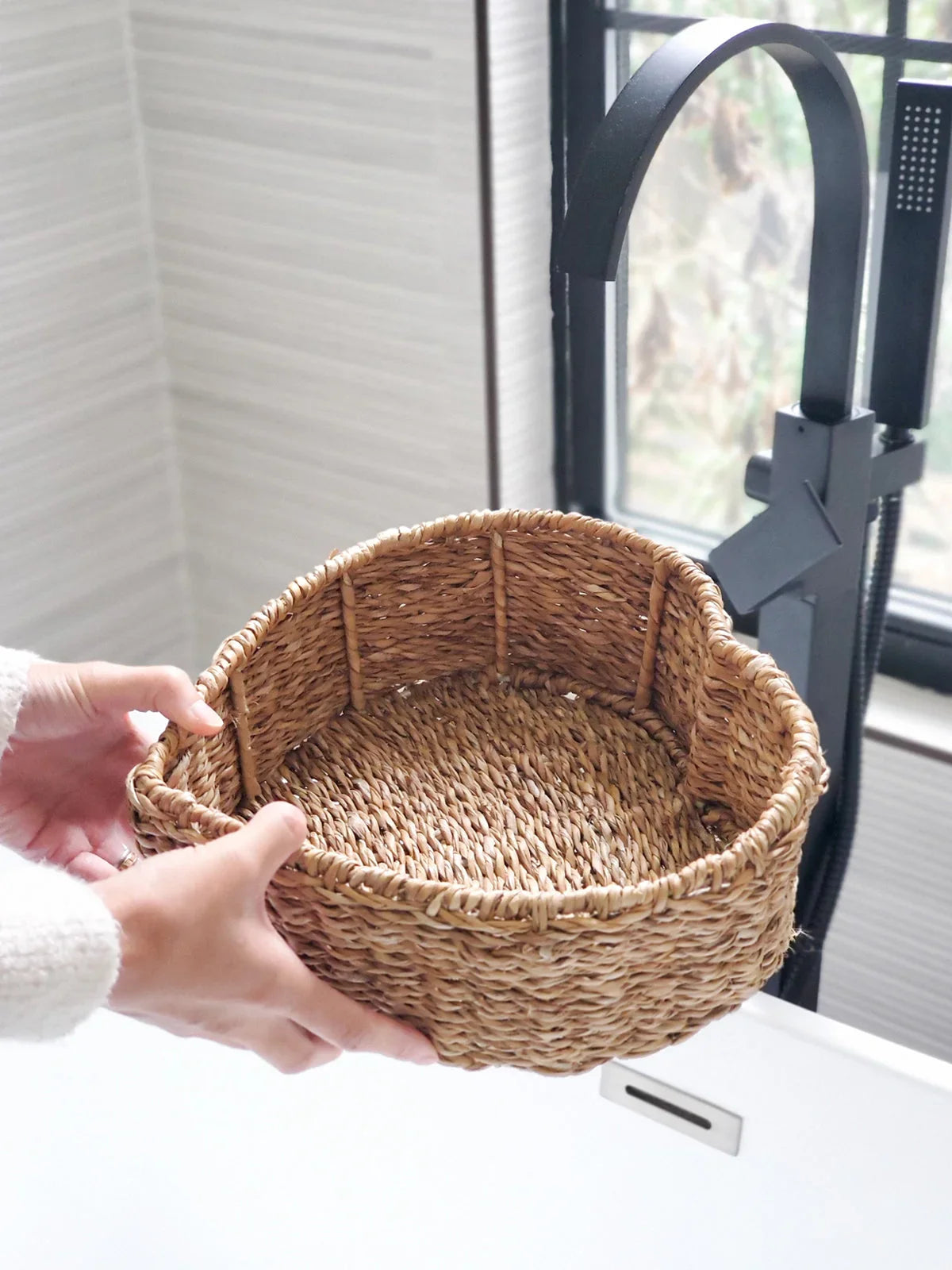 Hand holding a heart-shaped woven wicker basket near a black kitchen faucet and white sink