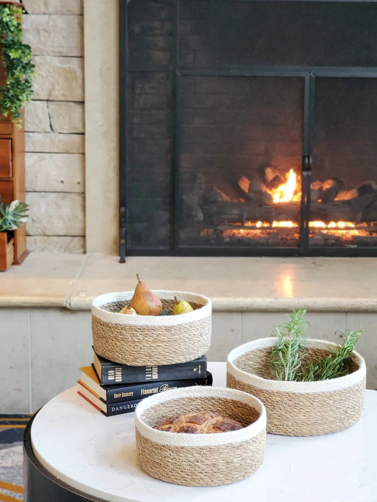 Set of three natural woven storage bowls on table by fireplace, styled with fruit, bread, and herbs at Qu!rk Boutique.