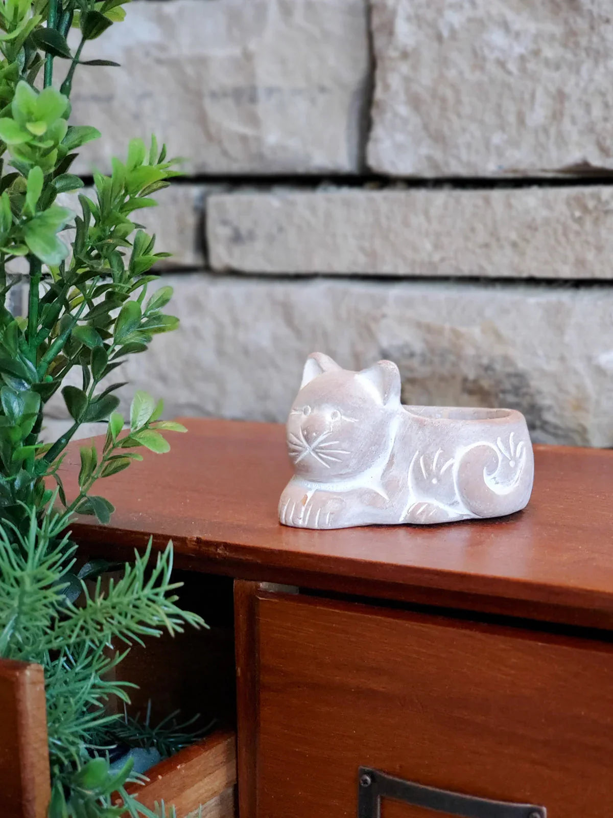 Terracotta cat-shaped candle holder on wooden table with green plants and stone wall background