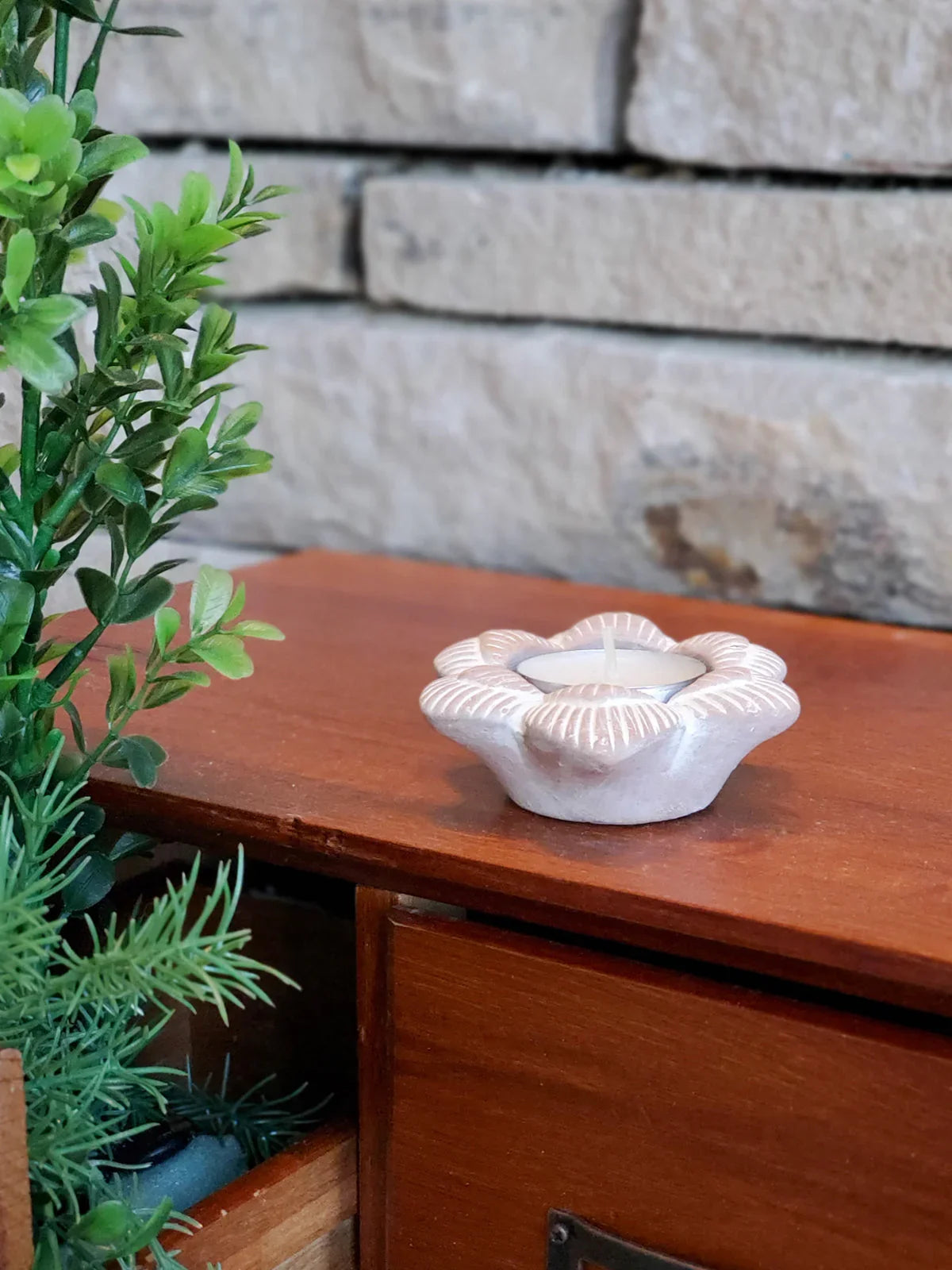 Terracotta flower-shaped candle holder with white tealight on wooden surface near green plant