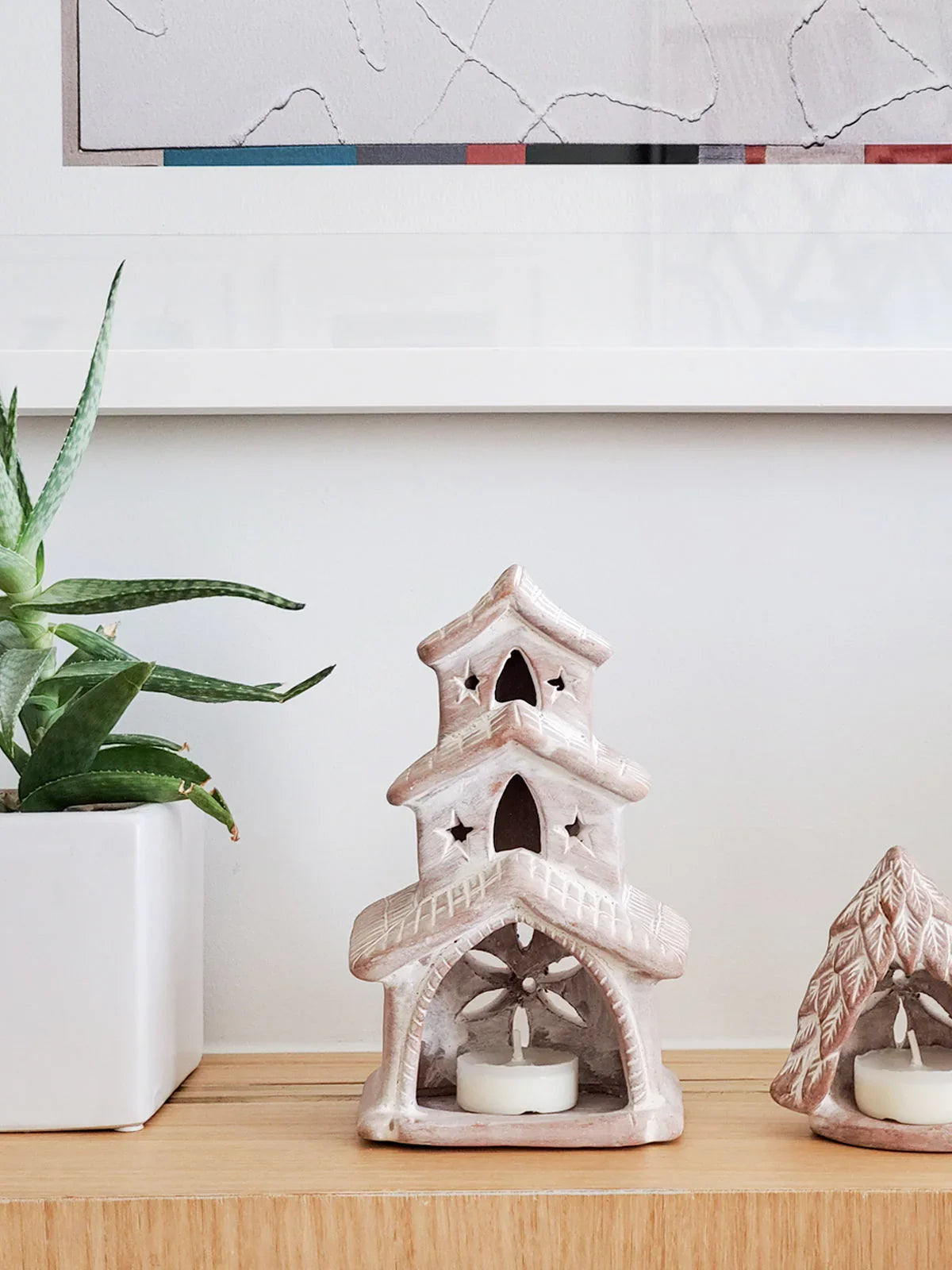 Terracotta house-shaped candle holder with star cutouts, wooden shelf with potted aloe plant