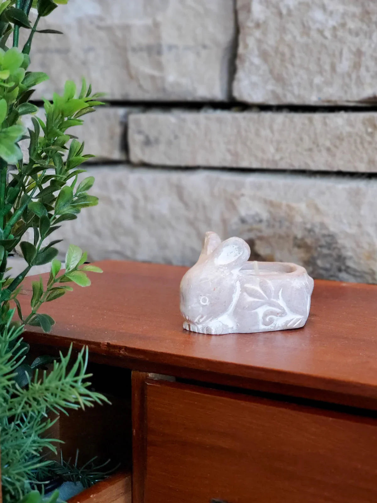 Terracotta rabbit-shaped candle holder on wooden surface with green plant and stone wall background