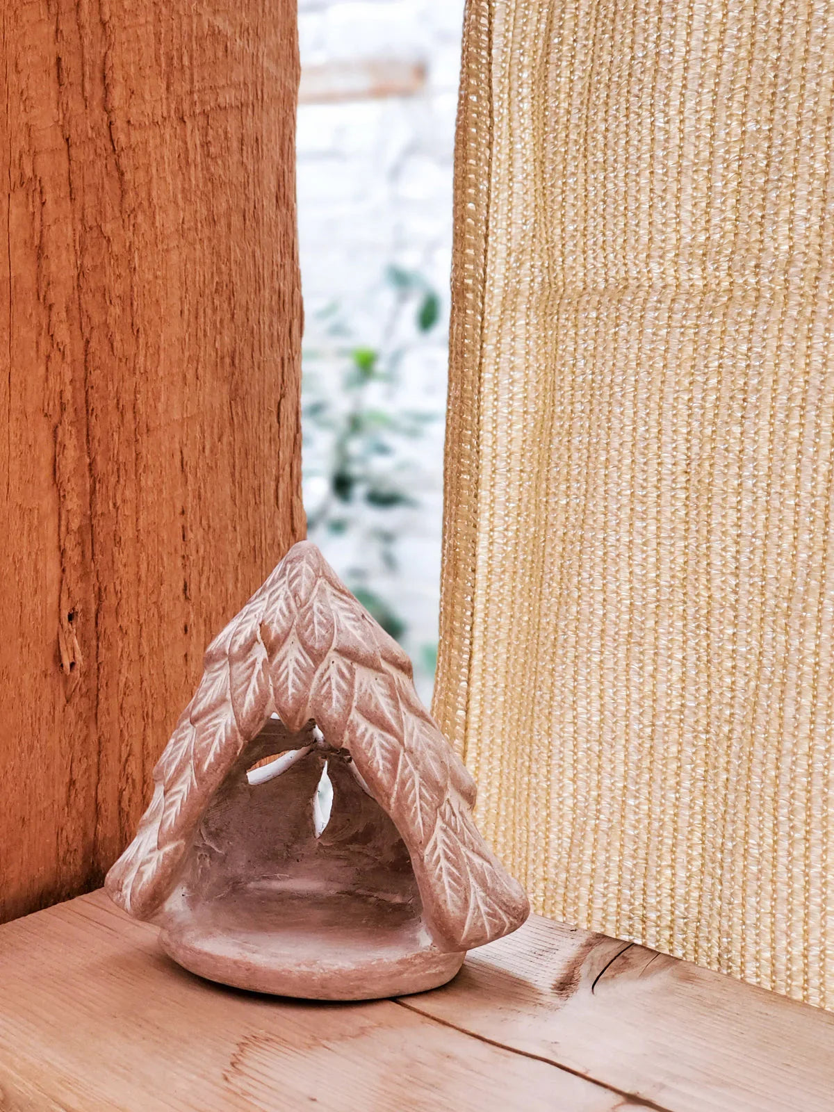 Terracotta tree-shaped candle holder with leaf pattern on wooden surface near window curtain