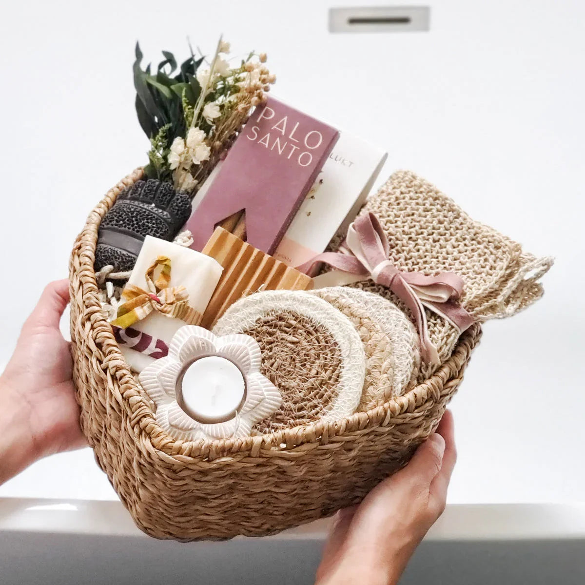 Hand holding woven spa gift basket with Palo Santo incense, candle, coasters, dried flowers, and knitted pouch