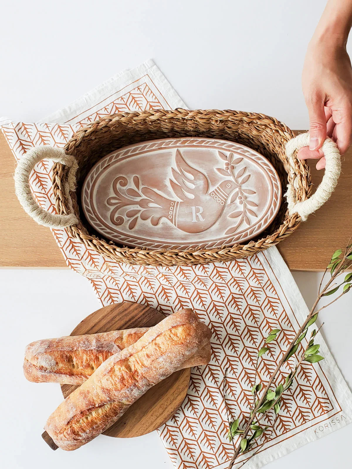 Woven bread warmer basket with rope handles holding an oval ceramic plate with bird and monogram R design next to fresh baguettes on wooden board