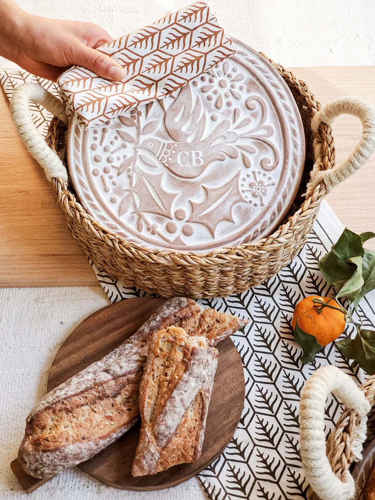 Woven bread warmer basket with decorative embossed lid featuring bird and foliage, side baguettes on wooden board