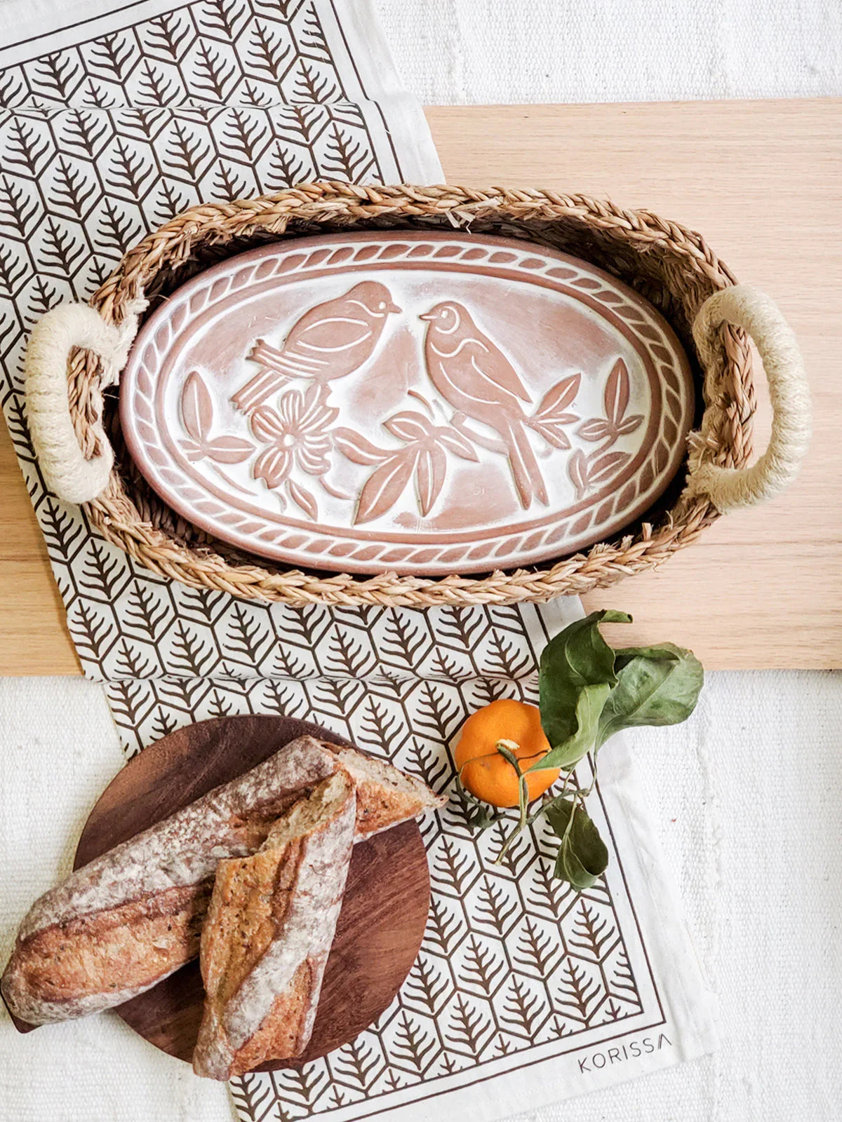 Oval ceramic bread warmer with bird design in woven basket on patterned cloth with bread and mandarin