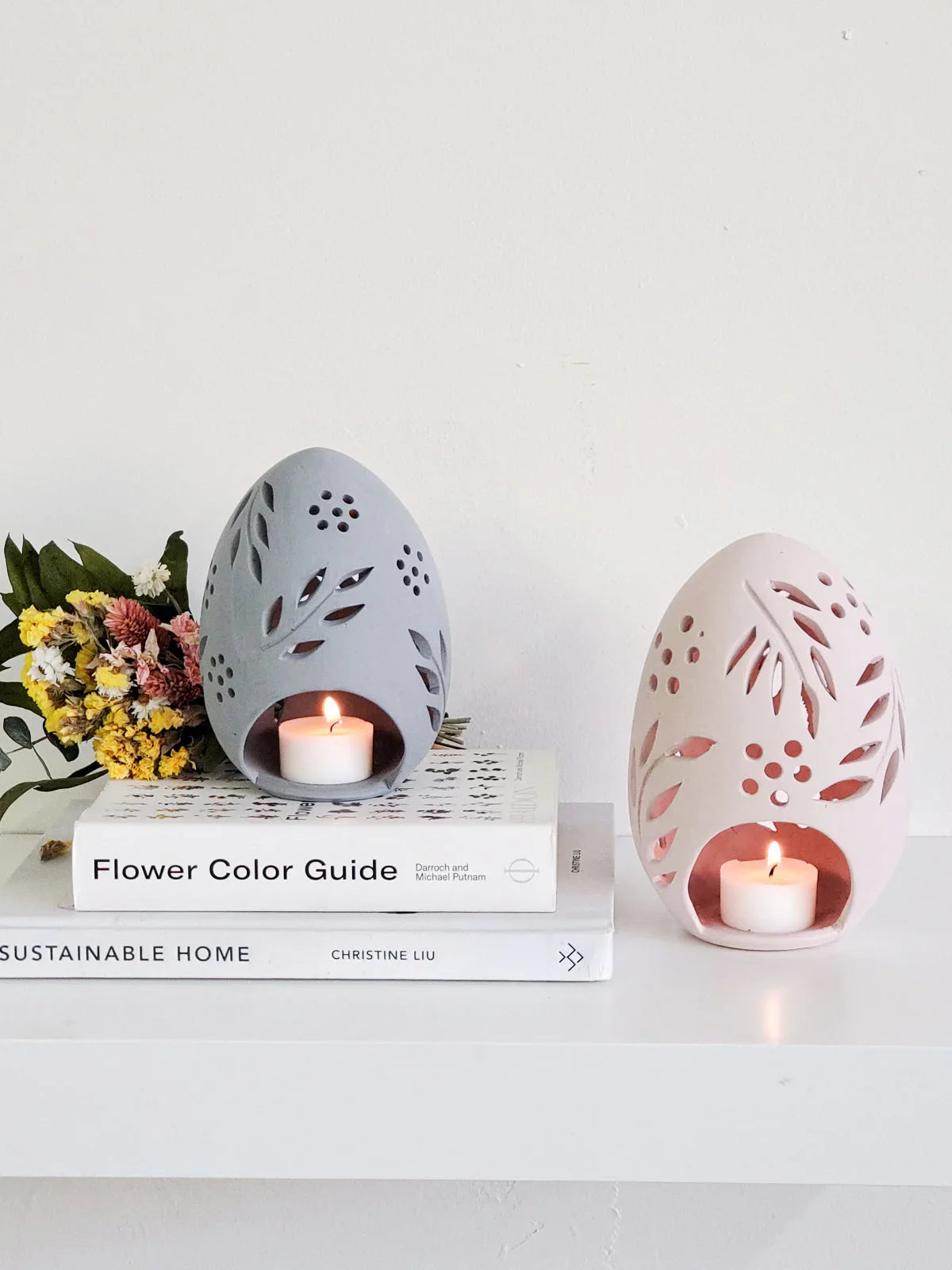 Terracotta egg-shaped candle lanterns with floral cutouts, one gray on books and one pink on shelf with burning candles