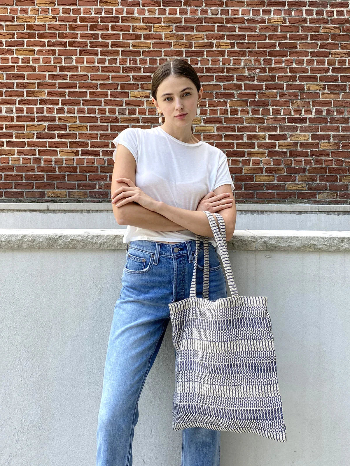 Woman in white t-shirt and jeans holding a blue and white tote bag, brick wall background