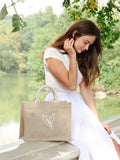 Woman in white dress sitting by lake with natural jute tote bag featuring leaf design