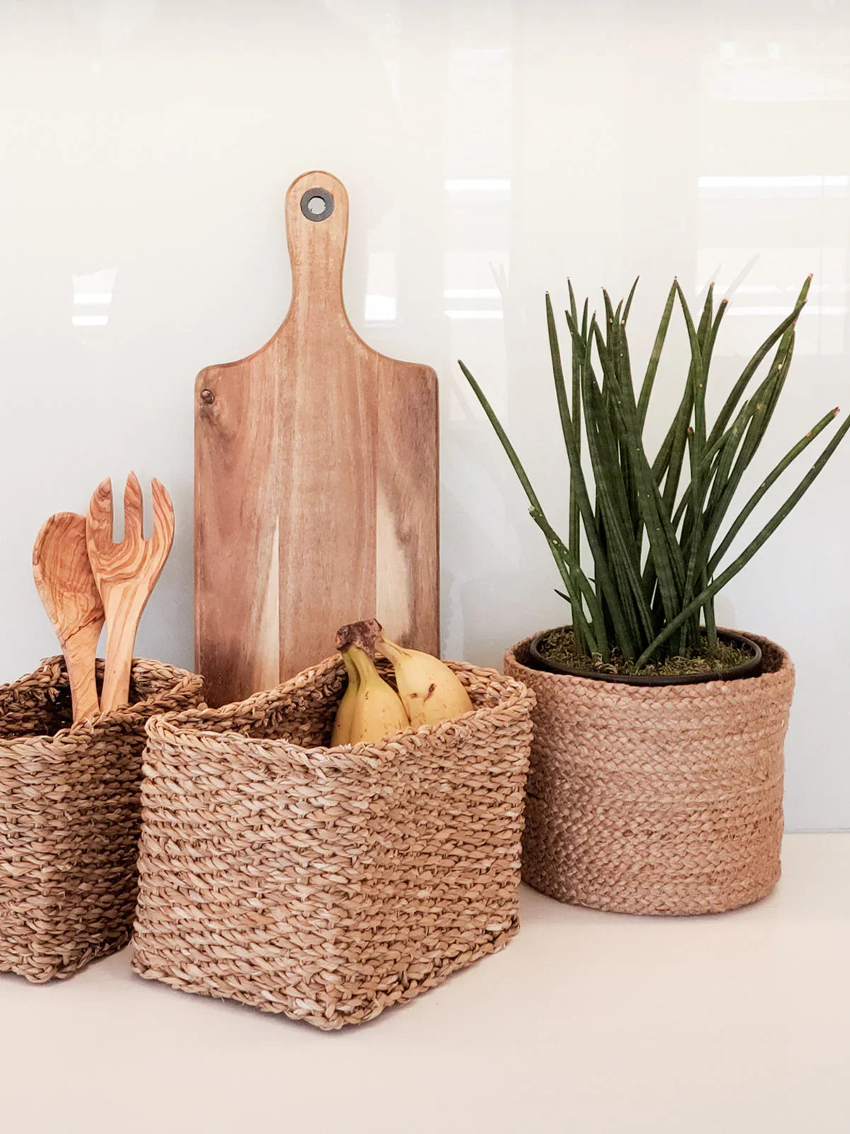 Natural woven baskets with wooden utensils, bananas, wooden cutting board, and potted green plant