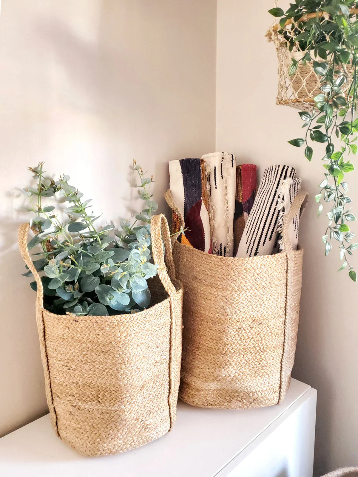 Natural woven baskets with handles, one holding eucalyptus, one holding textiles, on a white shelf near hanging greenery at Qu!rk Boutique.