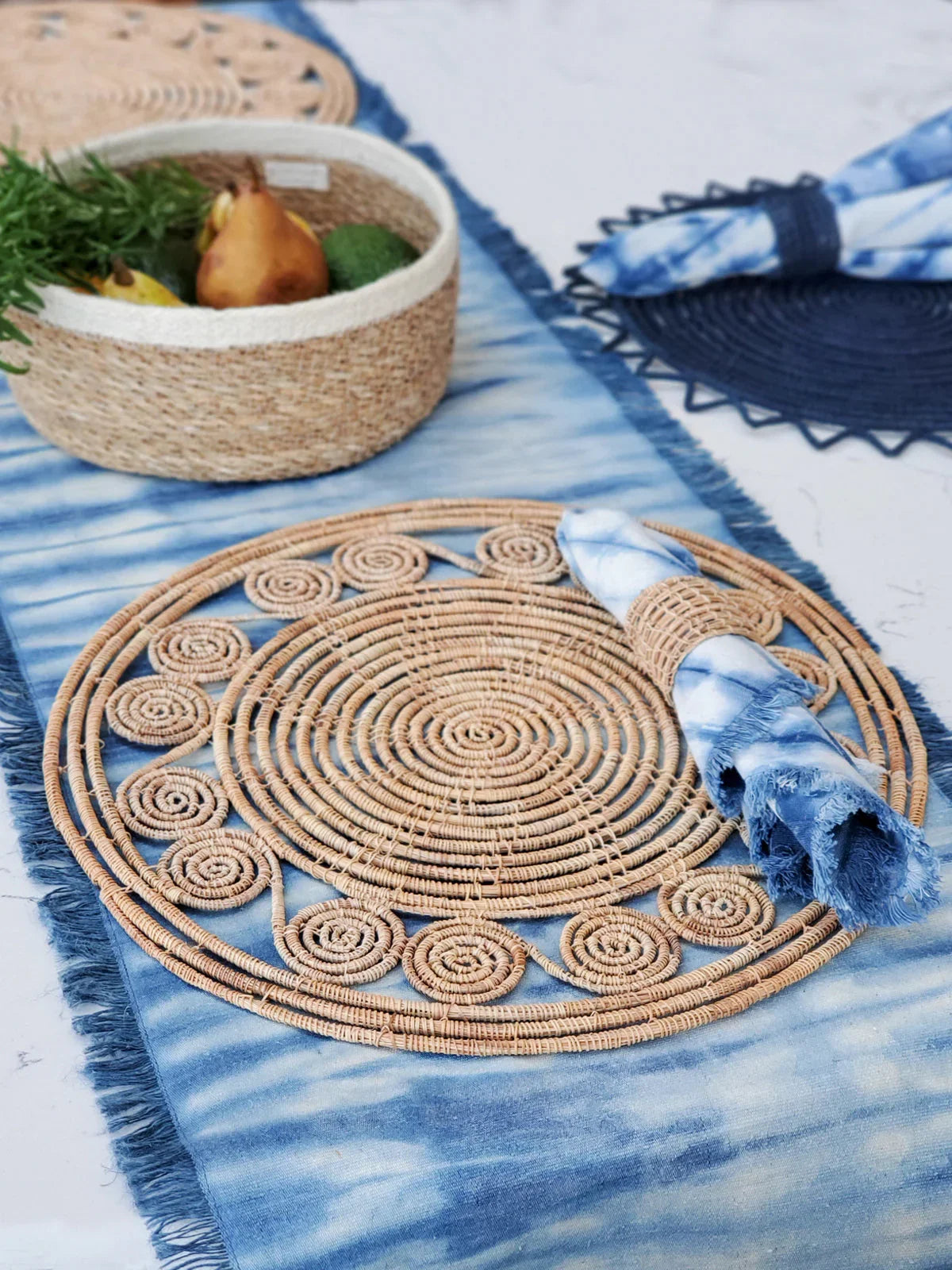 Natural woven rattan placemat with blue tie-dye napkin and a matching napkin ring on a blue table runner