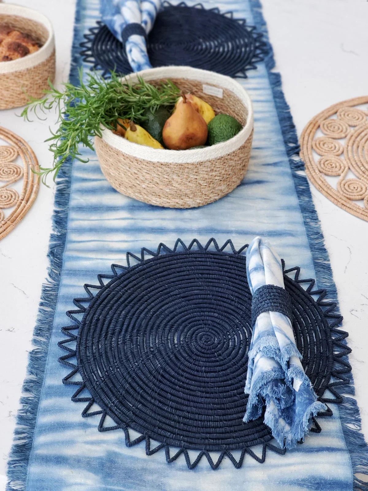 Table setting with navy woven placemat, blue tie-dye napkin, and woven basket holding pears and avocados on blue runner