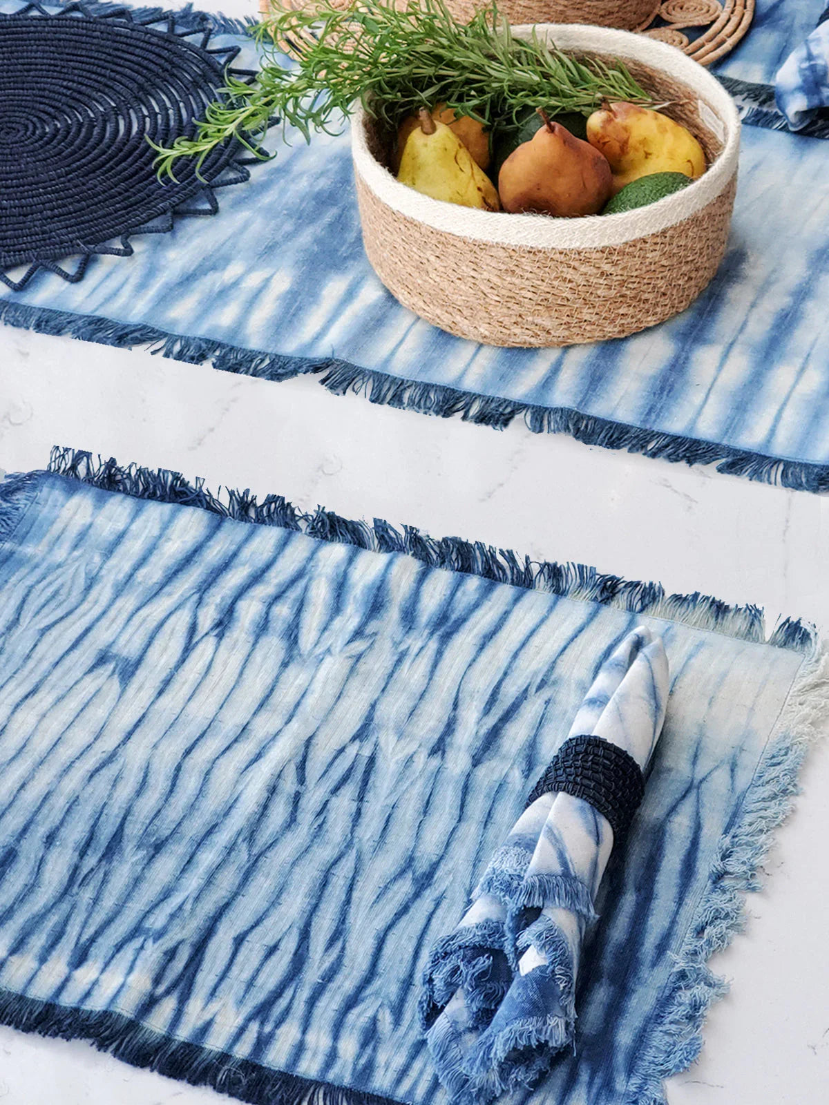 Blue tie-dye cotton placemats with fringed edges on a white surface, accompanied by matching napkin and woven basket with pears