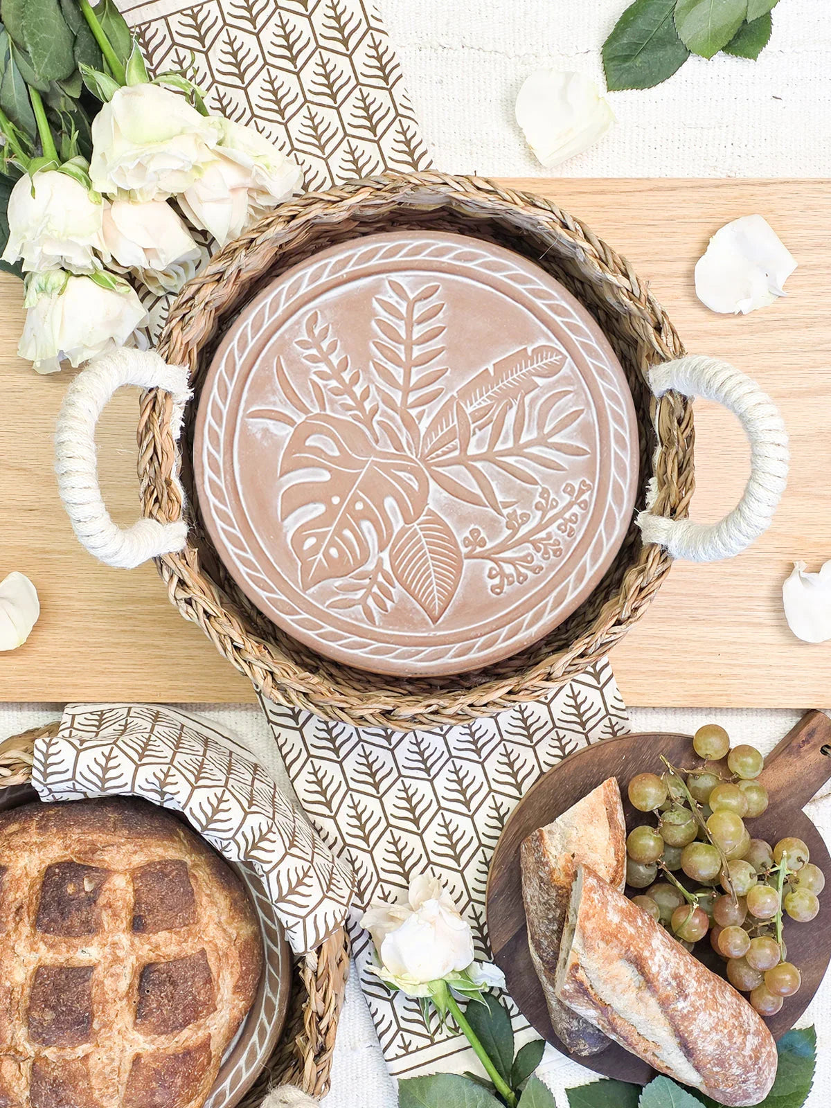 Woven basket with a decorative clay plate, fresh bread, grapes, and white roses on wooden board