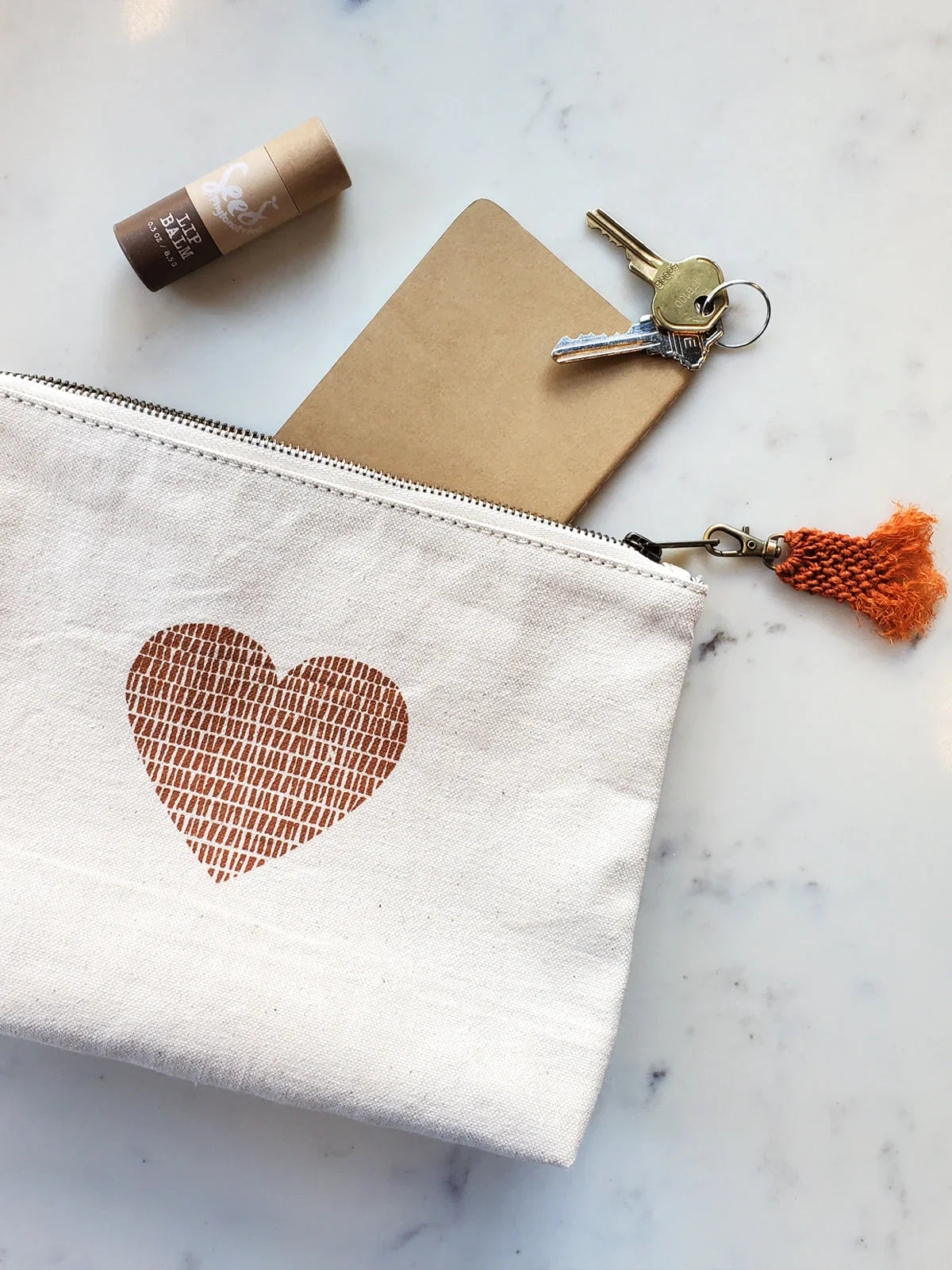 Canvas pouch with brown heart print, notebook, keys, and lip balm on white marble surface