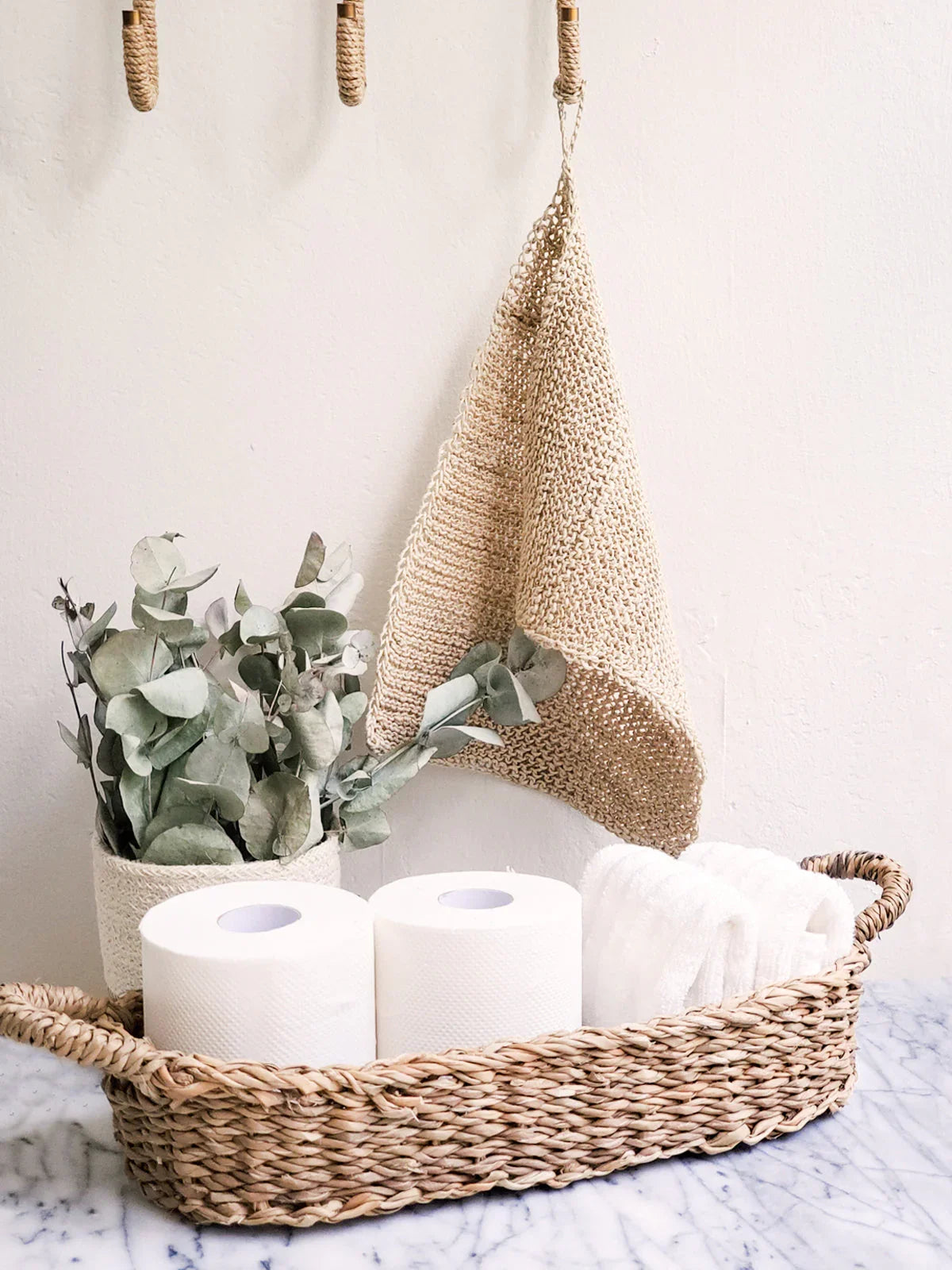 Woven natural handle bread basket holding two toilet paper rolls and white rolled towels on marble surface