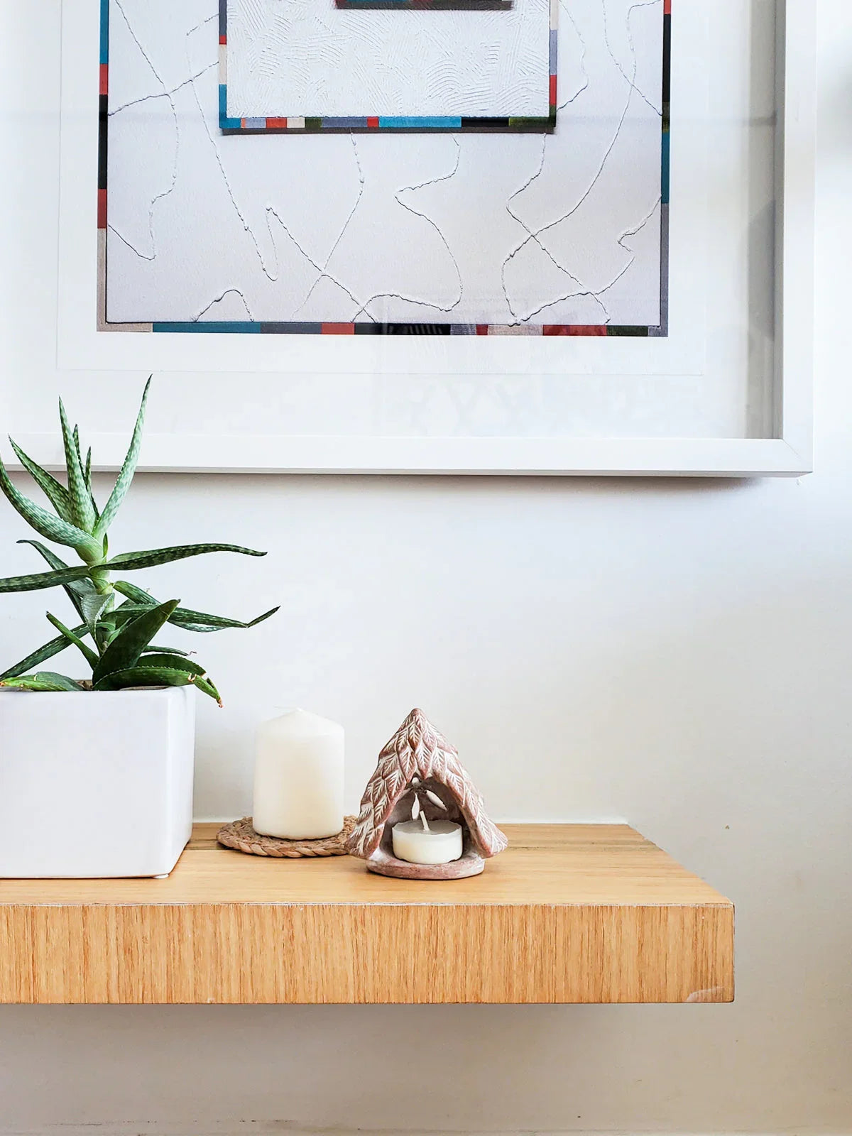 Terracotta tree-shaped candle holder with small white candle, aloe plant in white pot, white pillar candle on wood shelf