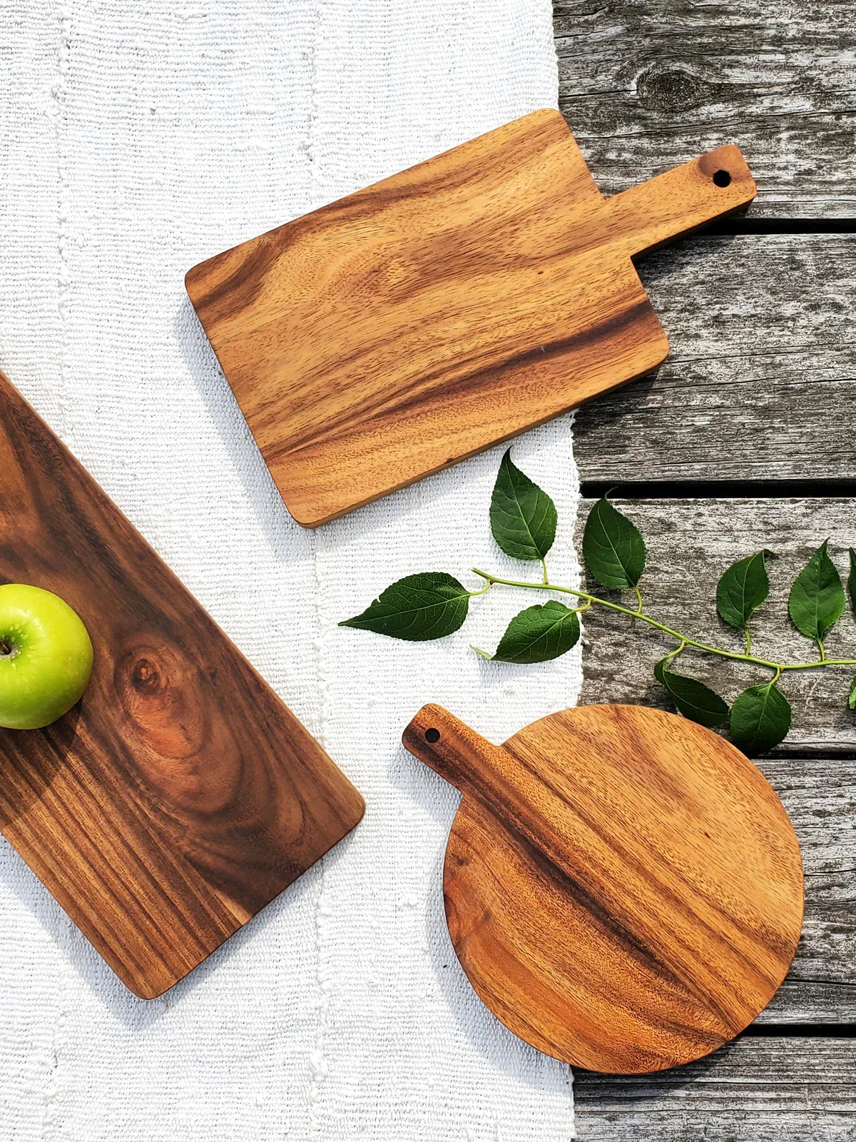 Two wooden serving boards and one wooden cutting board on white cloth and weathered wood surface with green apple and leaves
