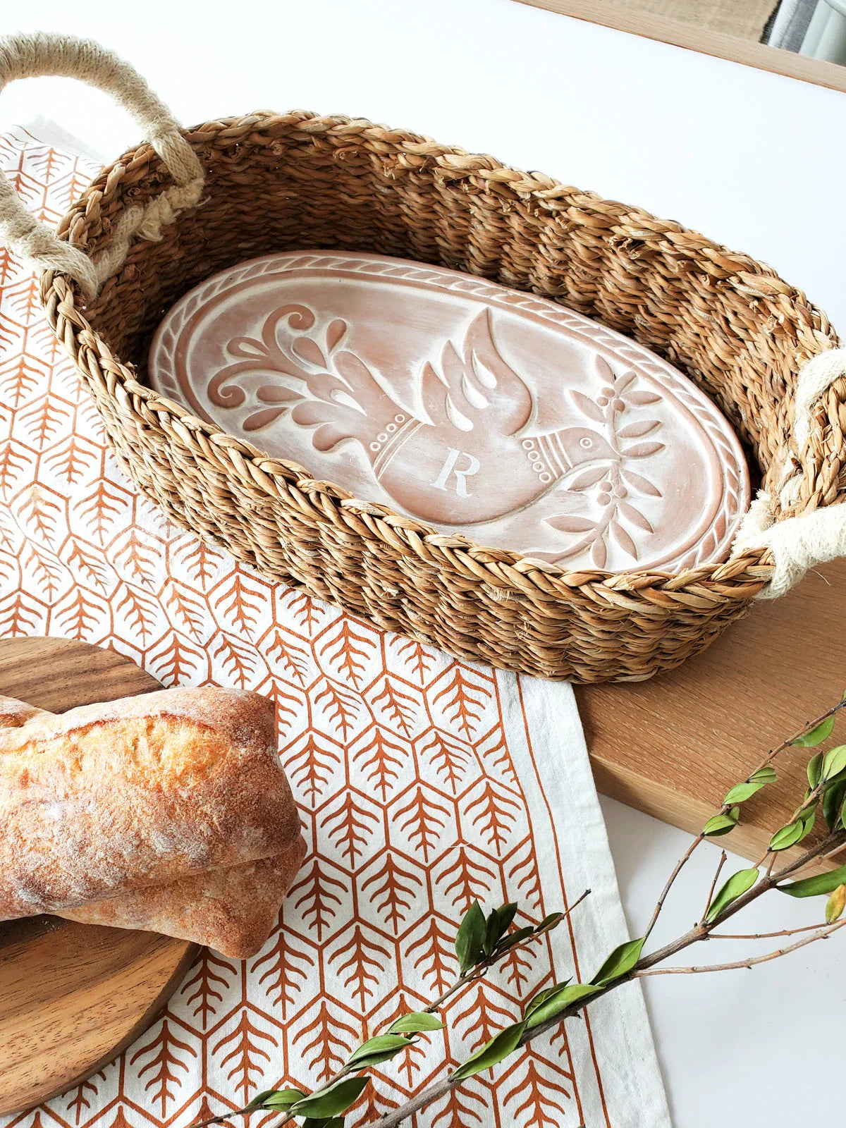 Woven bread warmer basket with monogrammed bird ceramic insert on patterned cloth next to rustic bread