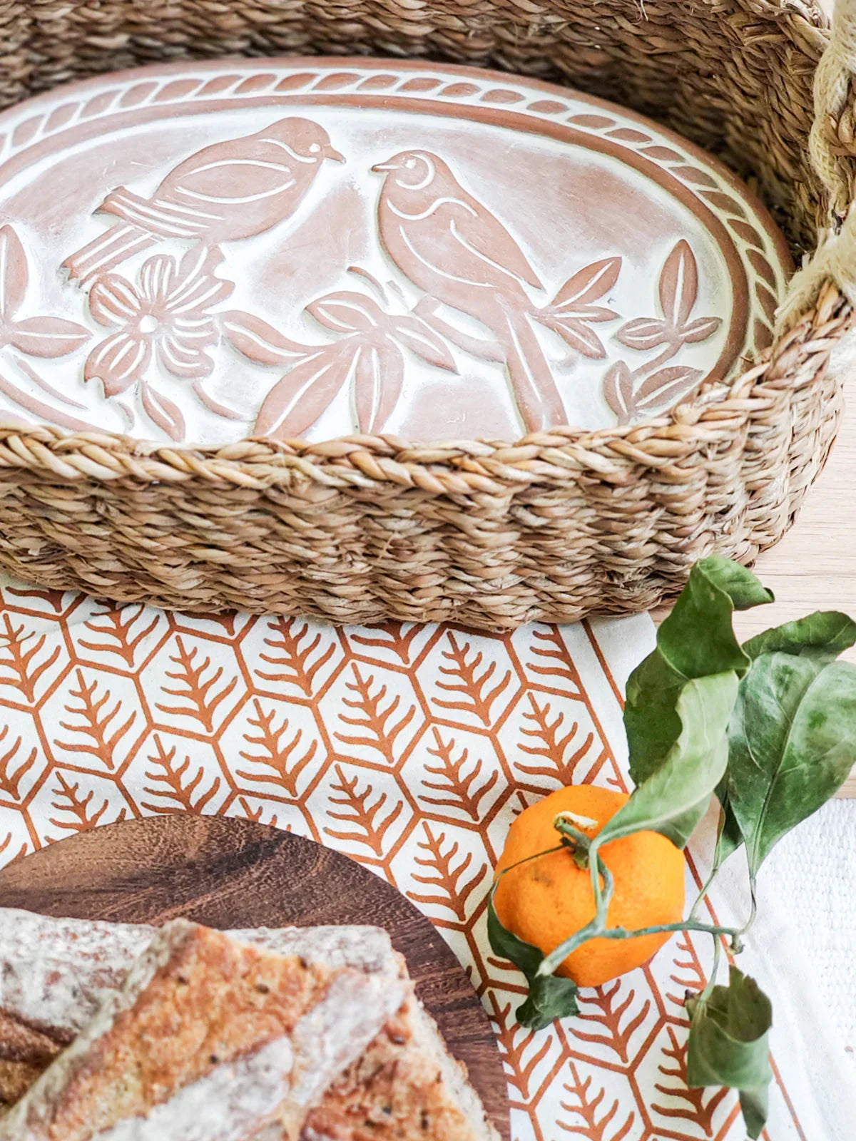 Oval woven bread warmer basket with bird design tray, wooden board with bread, and orange on patterned cloth