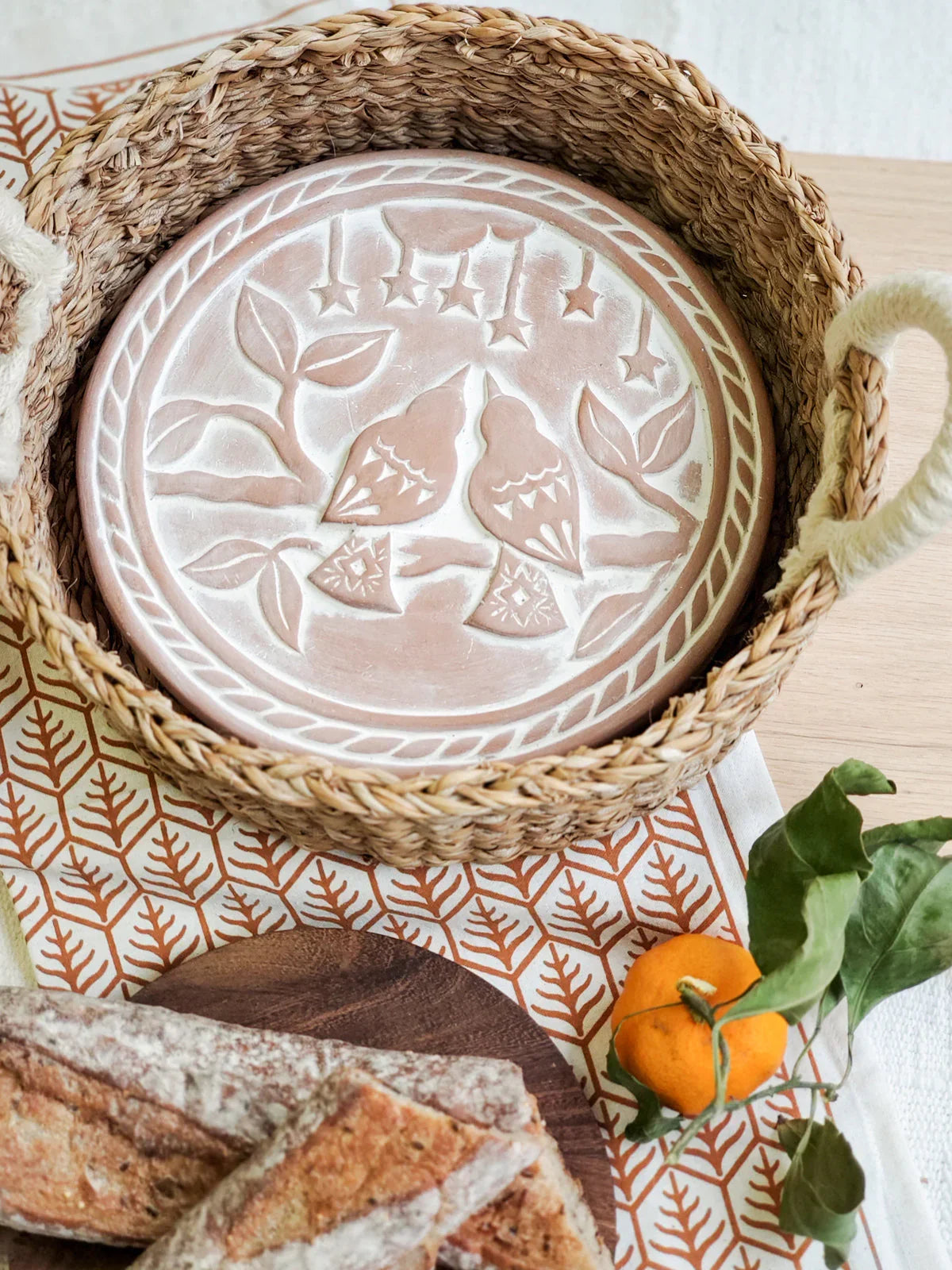 Round ceramic bread warmer with lovebird design inside woven basket on patterned cloth with bread and mandarin