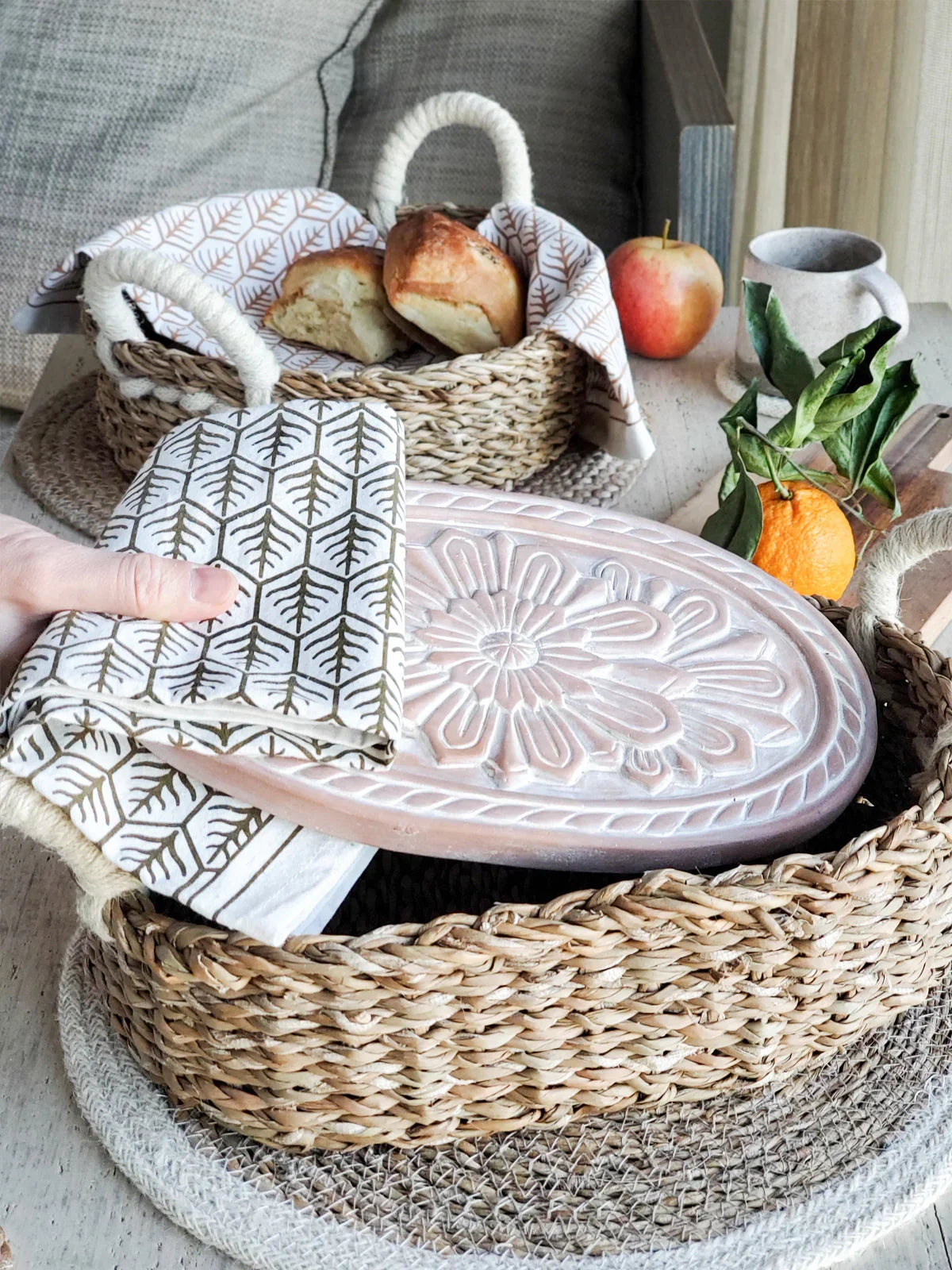 Hand holding patterned tea towel over pink floral ceramic bread warmer in woven basket with bread, apple, orange, and cup