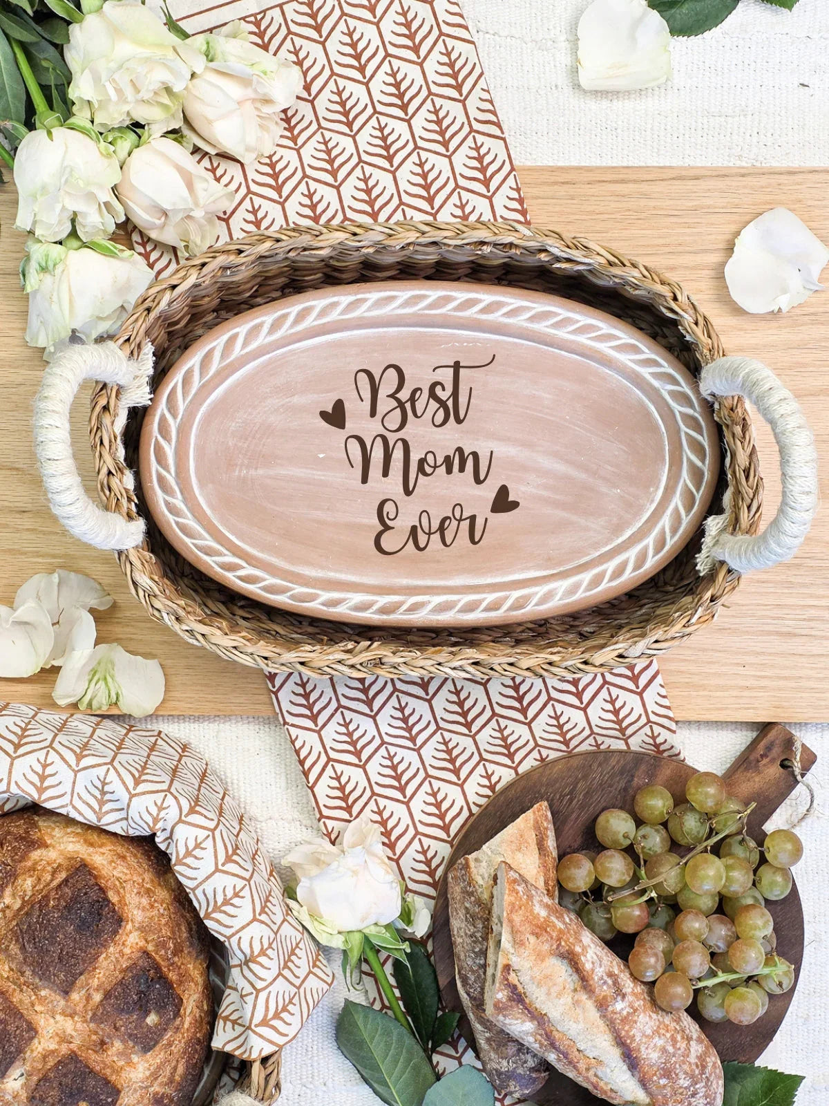 Woven basket with oval tray saying Best Mom Ever, surrounded by bread, grapes, and white roses