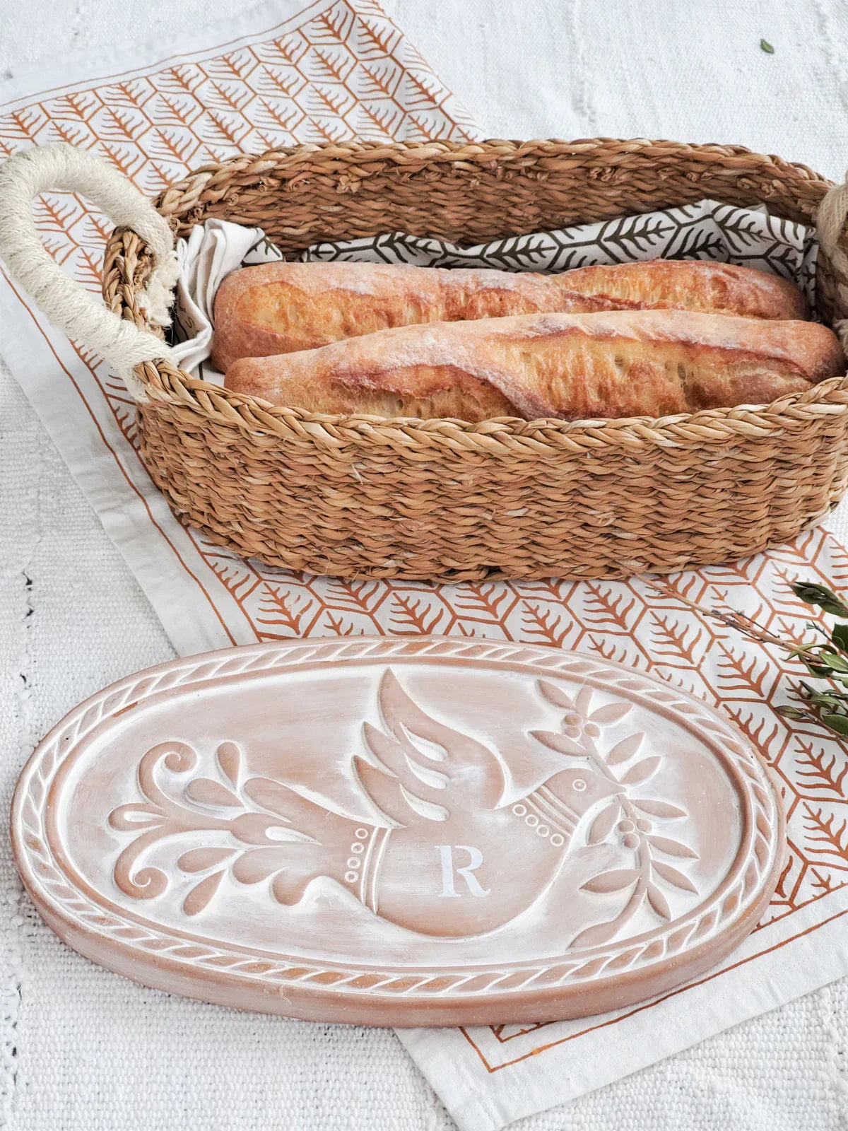 Oval bread warmer with carved dove design and letter R next to wicker basket holding baguettes