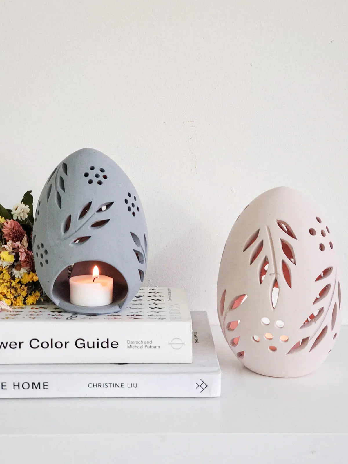 Terracotta egg-shaped candle lanterns with leaf cutouts, one grey lit on books, one pale pink