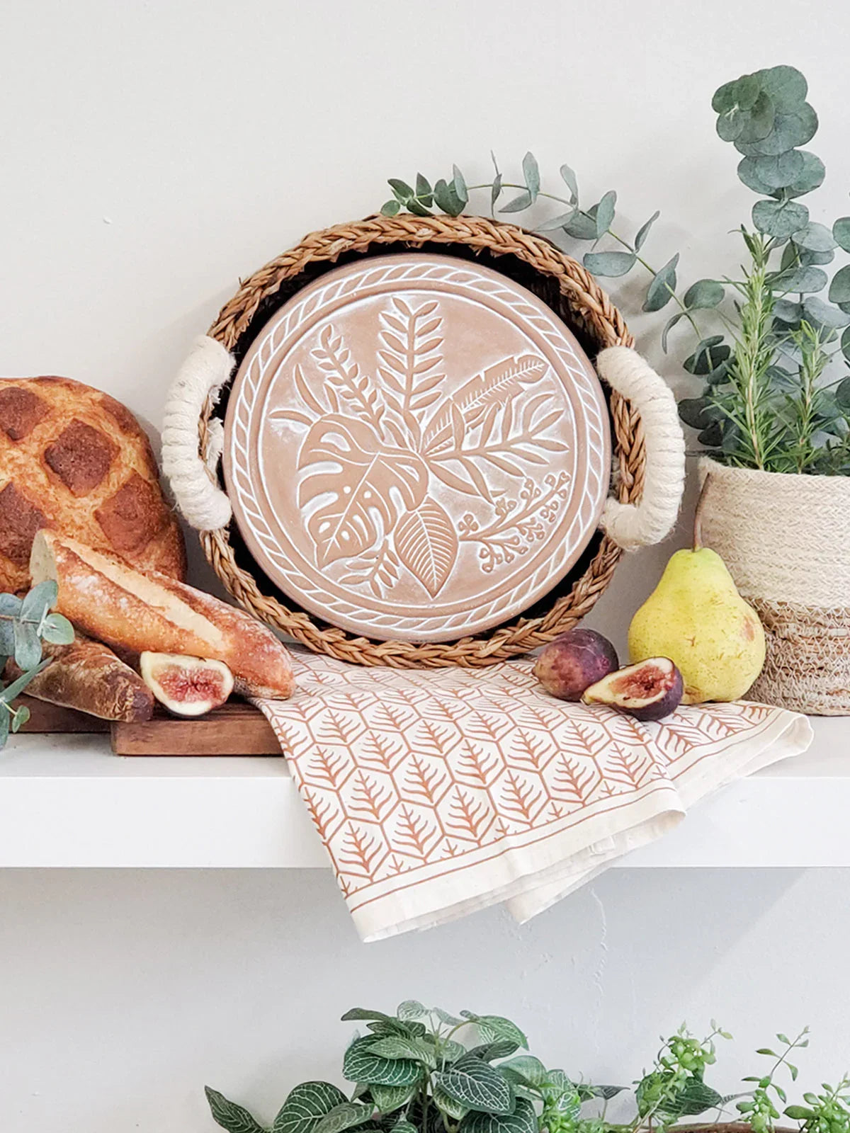 Woven basket with embossed plant design plate, patterned tea towel, bread, figs, pear, and potted plants on white shelf