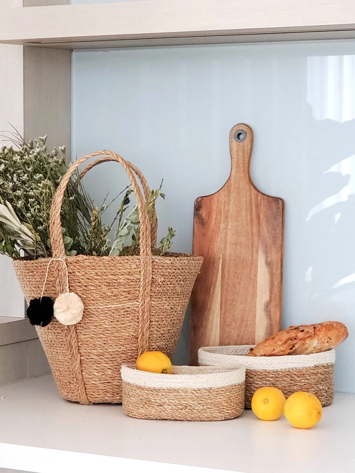 Set of three woven seagrass baskets on a kitchen counter with lemons and bread, styled with a wooden cutting board, at Qu!rk Boutique & Gift Shop.