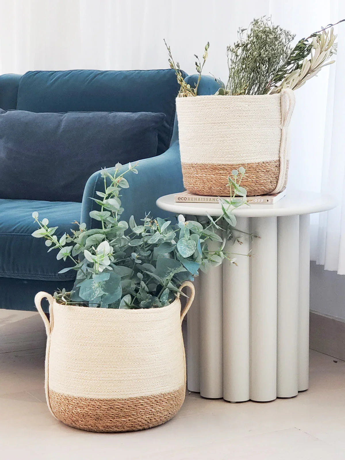 Two woven storage baskets with plants, one on a white modern round side table and one on the floor near a blue sofa