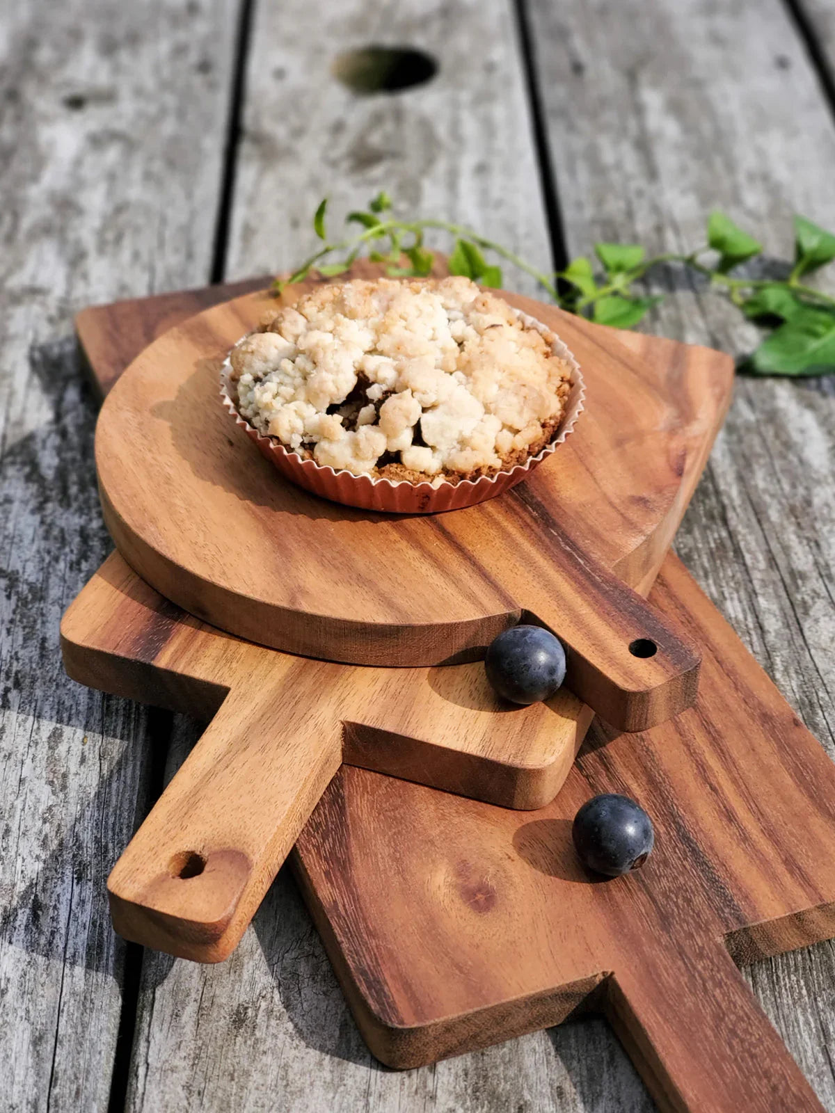 Crumb-topped dessert on small round wooden serving board with two blueberries on rustic wooden table