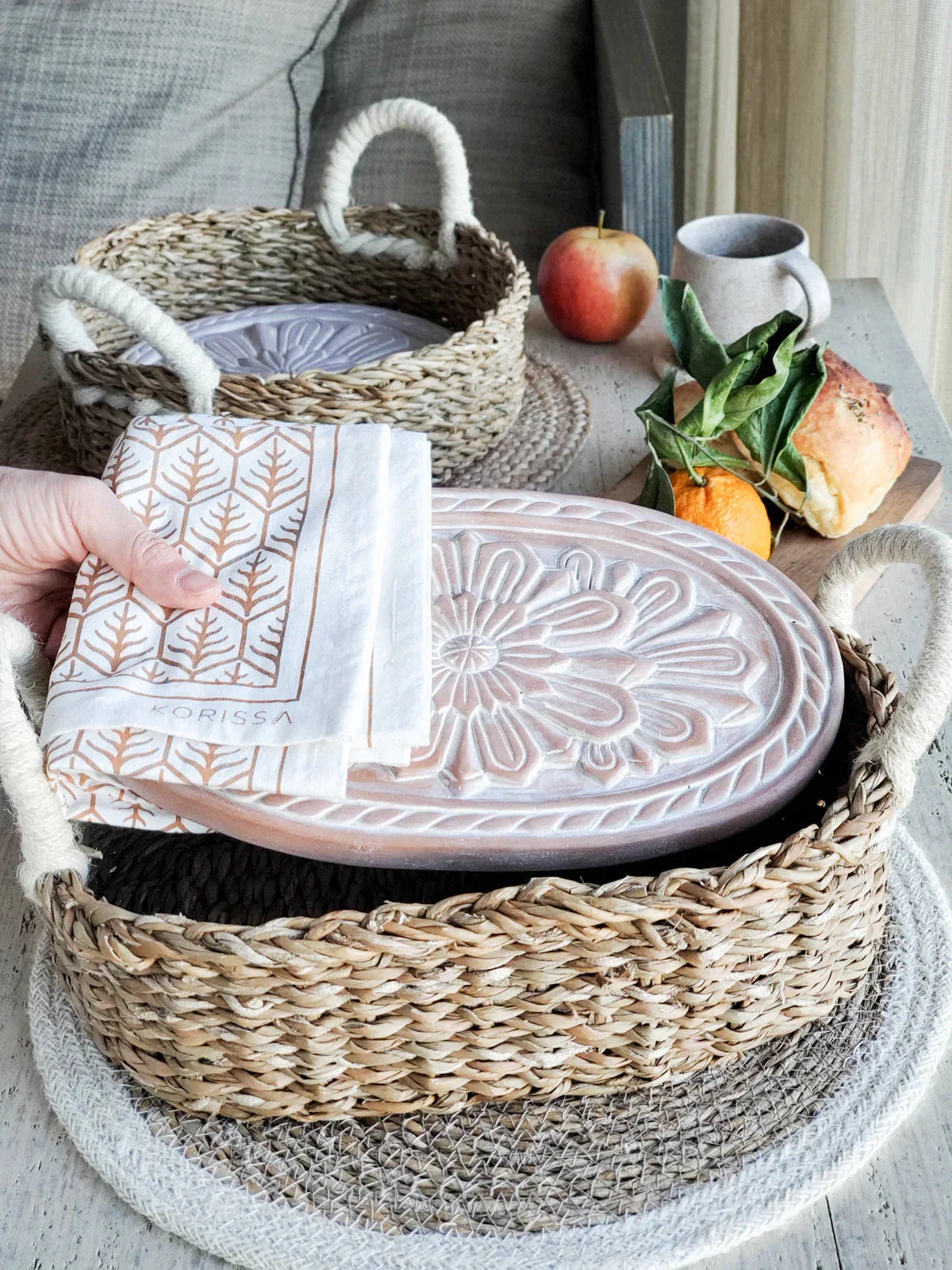 Woven bread warmer basket with floral embossed ceramic lid and patterned tea towel on wooden table