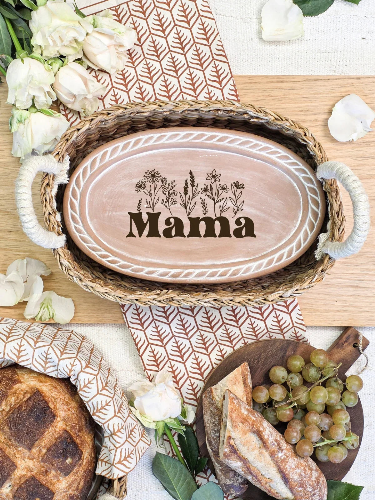 Oval bread warmer with floral design and text Mama in wicker tray surrounded by bread, grapes, and white roses