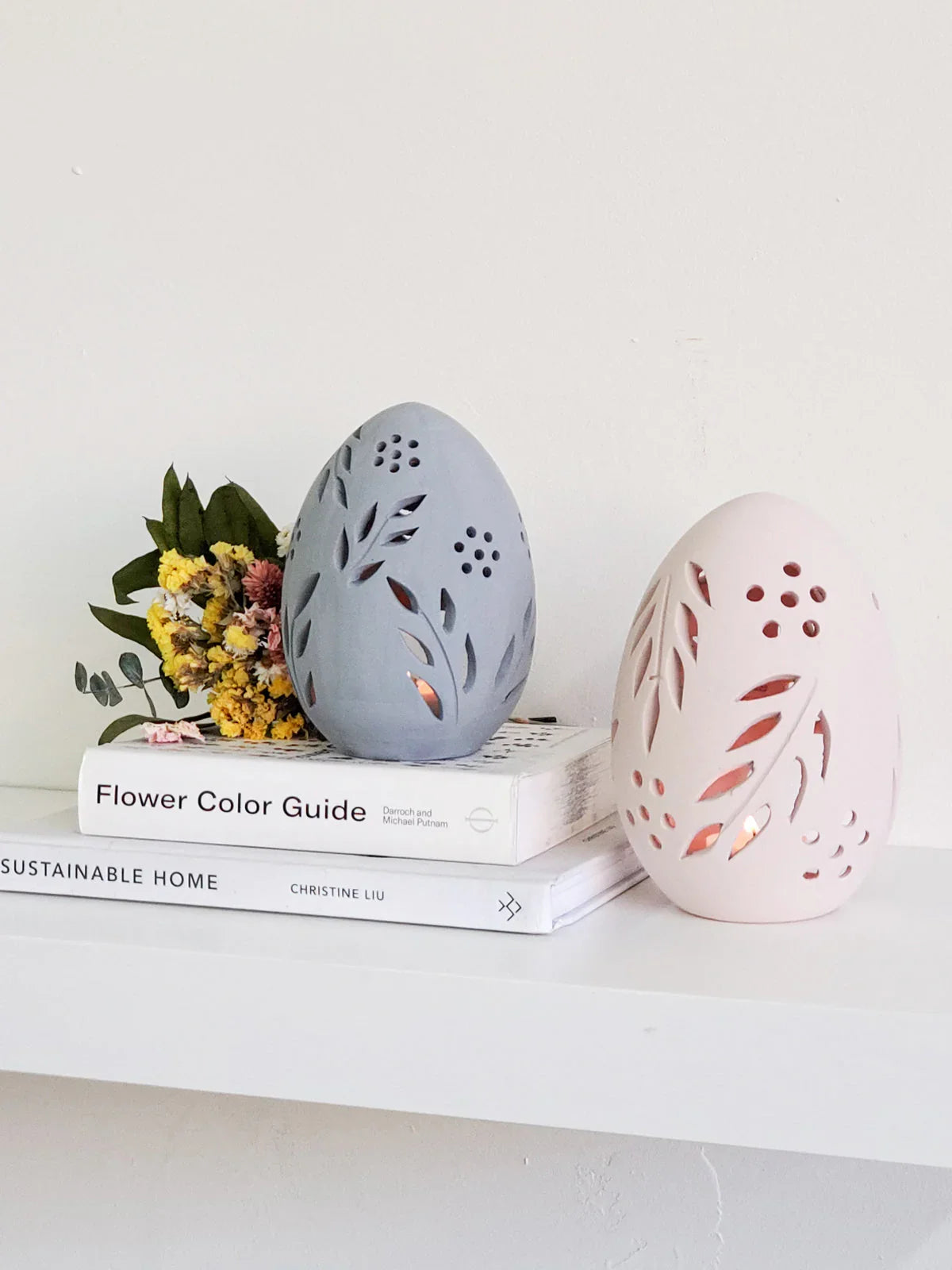 Decorative terracotta egg candle lanterns with floral cutouts on white shelf with books and dried flowers