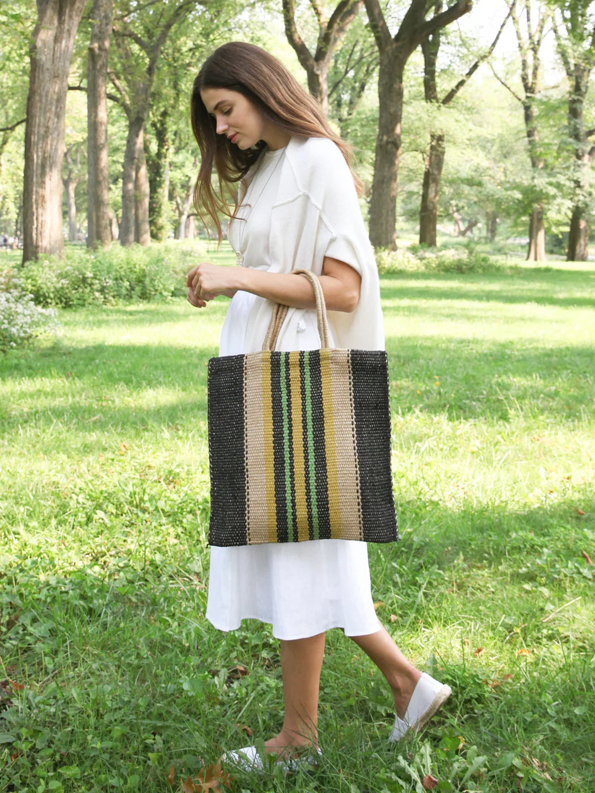 Woman in white dress carrying striped jute tote bag walking in sunlit park with green grass and trees