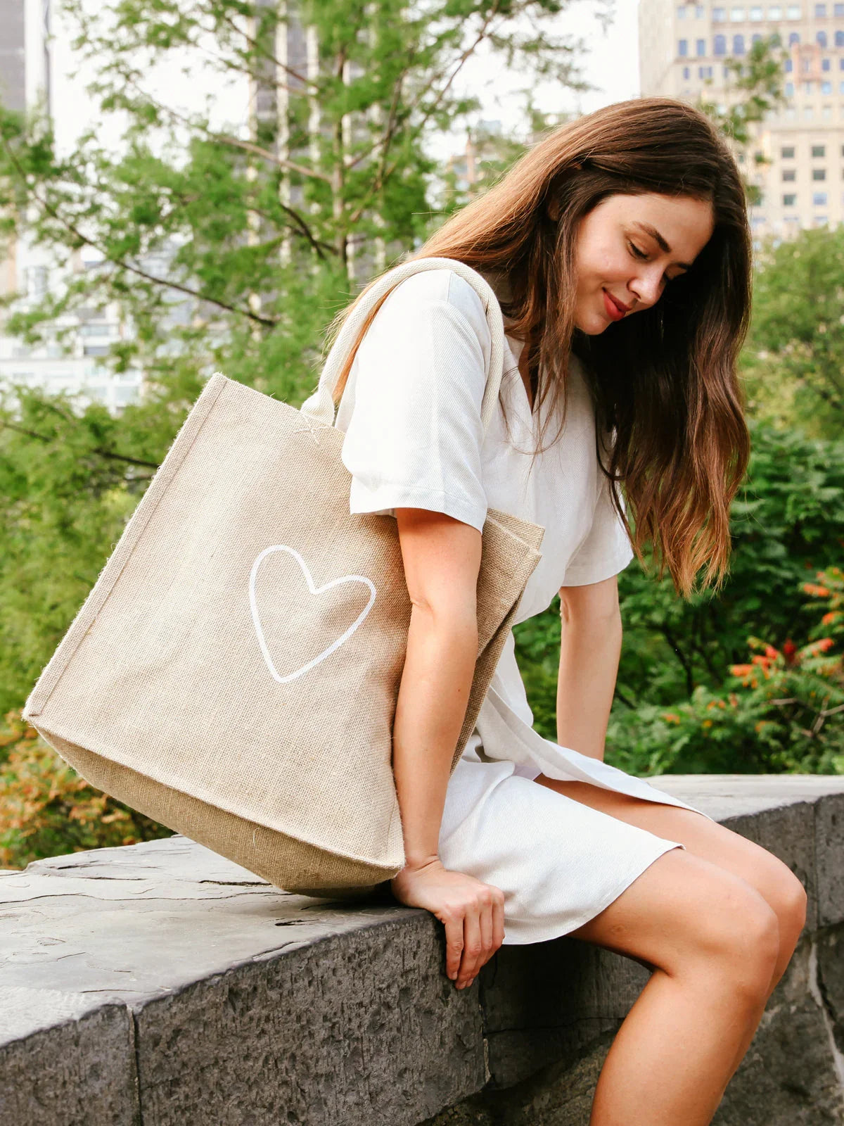Woman in white dress sitting on stone ledge outdoors with beige tote bag featuring white heart design
