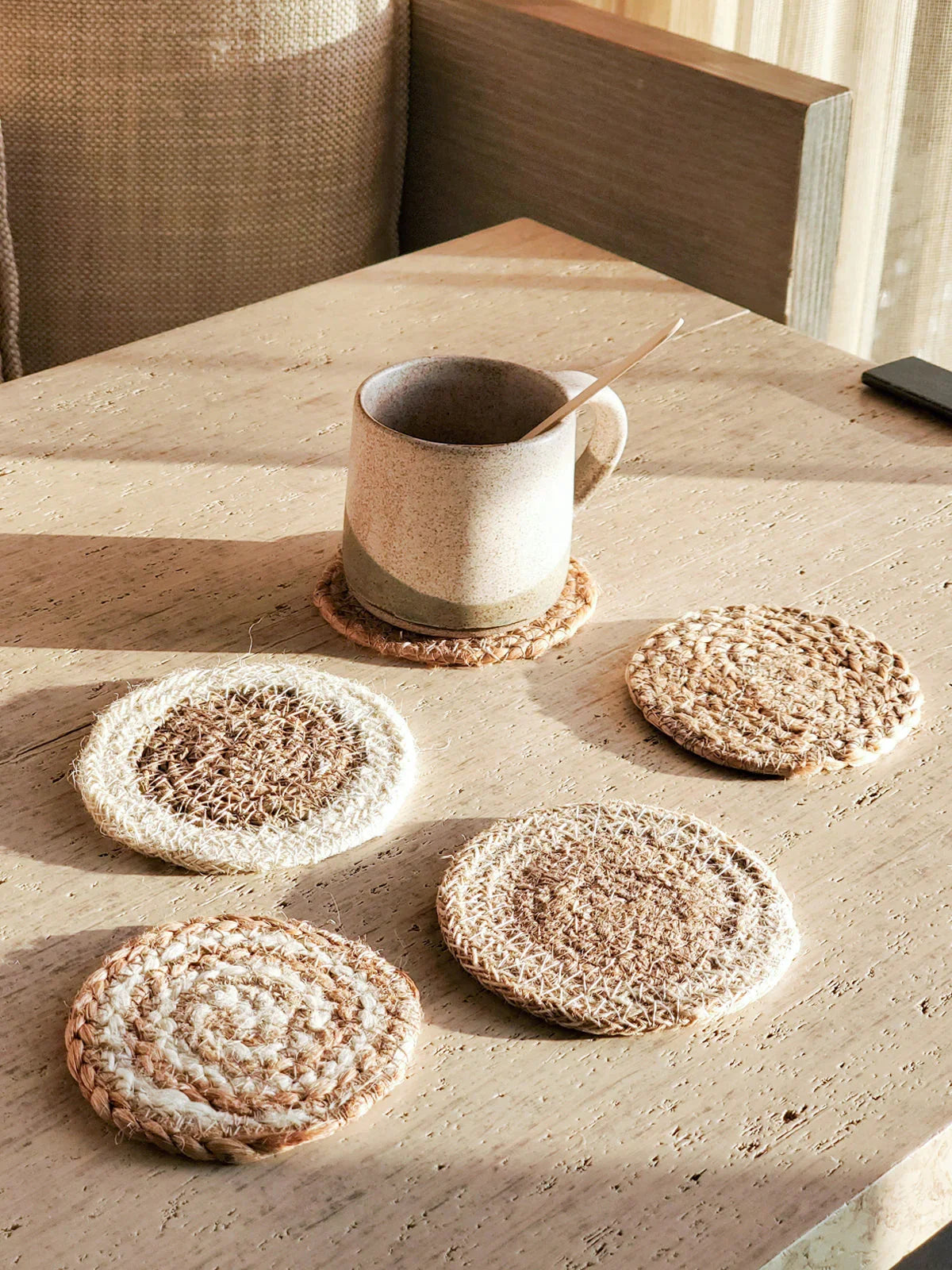 Ceramic mug with wooden spoon on woven coaster surrounded by four handmade jute coasters on wooden table