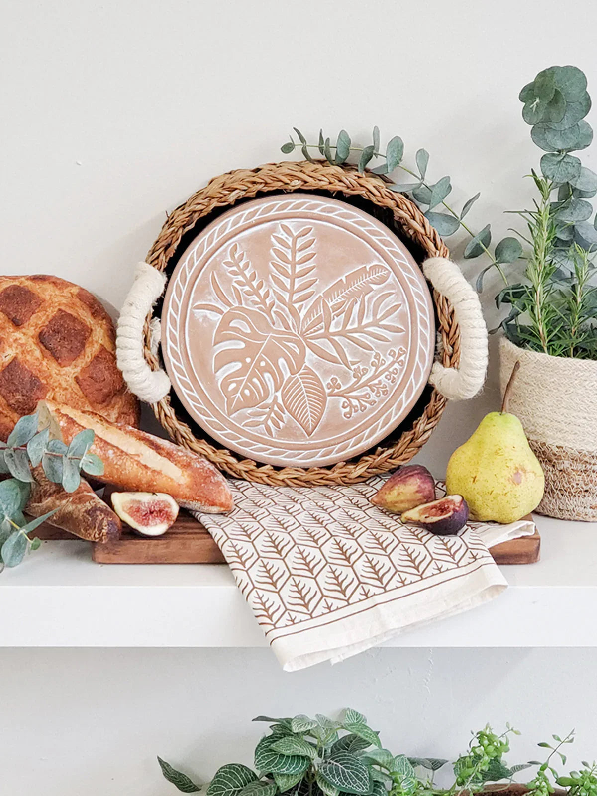 Woven basket with botanical print tray, patterned tea towel, fresh bread, figs, pear, and potted greenery on white shelf