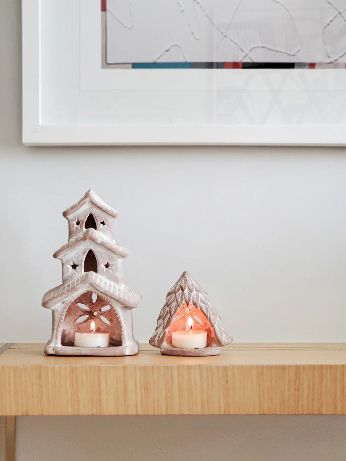 Two terracotta candle holders shaped like a multi-tiered house and a pine tree on a wooden shelf