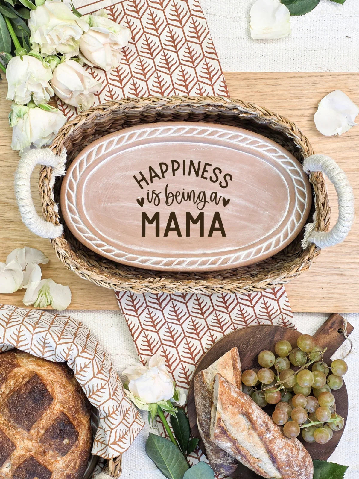 Oval wooden bread warmer with 'Happiness is being a Mama' text inside wicker basket on floral cloth with bread, grapes, and white roses