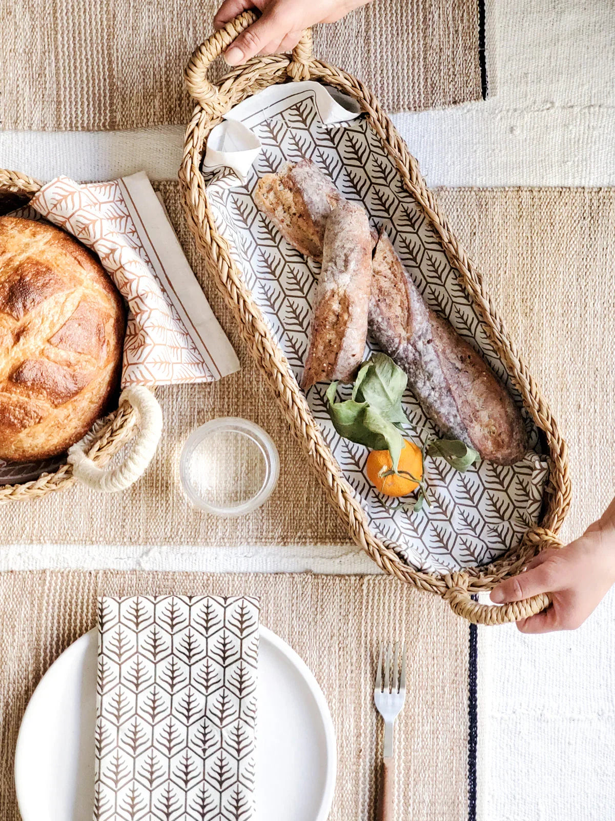 Oval woven bread basket with two baguettes and a small orange on patterned cloth, on a rustic table setting