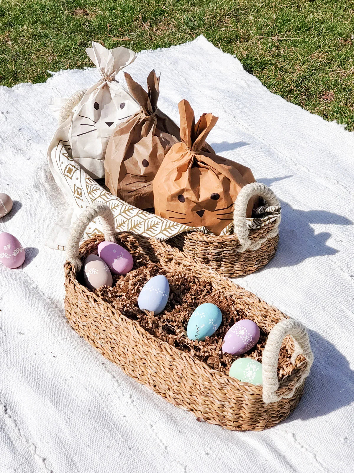 Two woven baskets on a white blanket outdoors; one basket holds cracked Easter eggs, the other holds three bunny-shaped paper bags