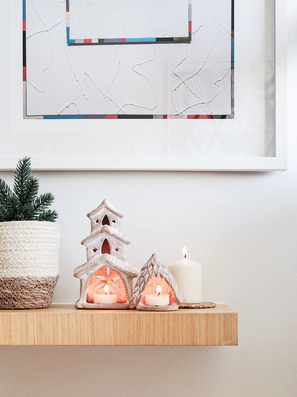 Terracotta house-shaped candle holders with lit candles on wooden shelf, next to white pillar candle and potted plant